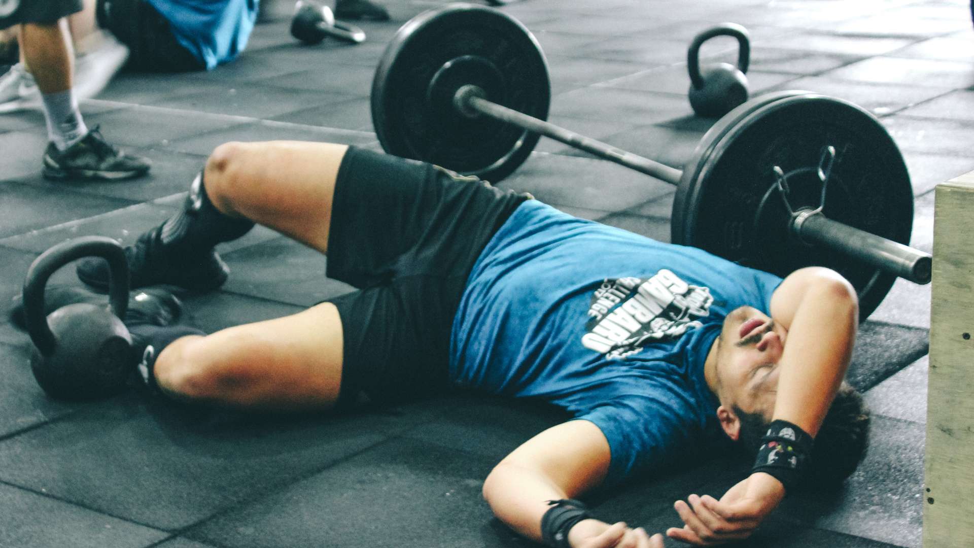 Bărbat întins epuizat pe podeaua sălii lângă o bară cu greutăți și kettlebell-uri, ilustrând oboseala și lipsa de energie