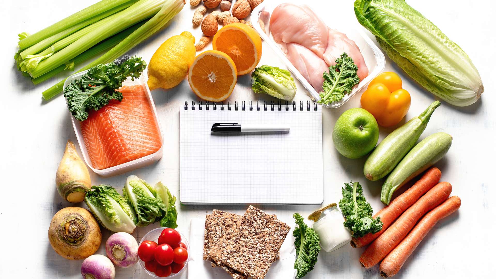 Aranjament văzut de sus cu alimente proaspete sărace în carbohidrați (țelină, pește, pui, legume, citrice, nuci) în jurul unui carnețel gol cu pix, pentru planificarea meniului