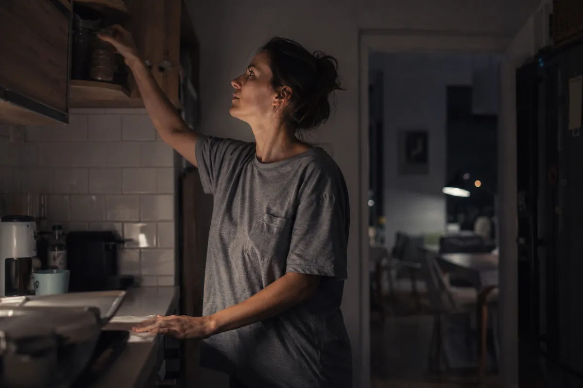 Femeie într-un tricou gri întinzându-se după un raft din bucătărie, noaptea, într-o bucătărie slab luminată